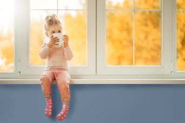 Little cute Kid Girl sitting by window holding cup of hot drink cocoa enjoying autumn forest...