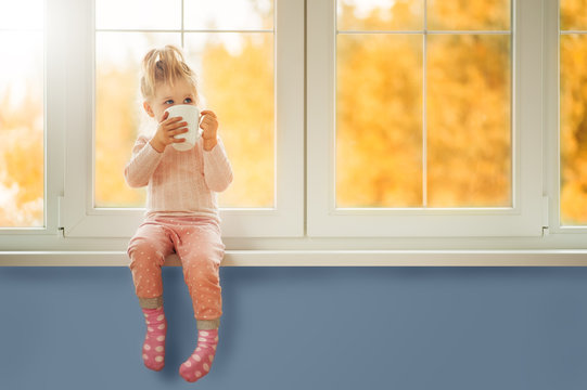 Little Cute Kid Girl Sitting By Window Holding Cup Of Hot Drink Cocoa Enjoying Autumn Forest Background. Season Beauty Fashion Children Concepts. Fall Season