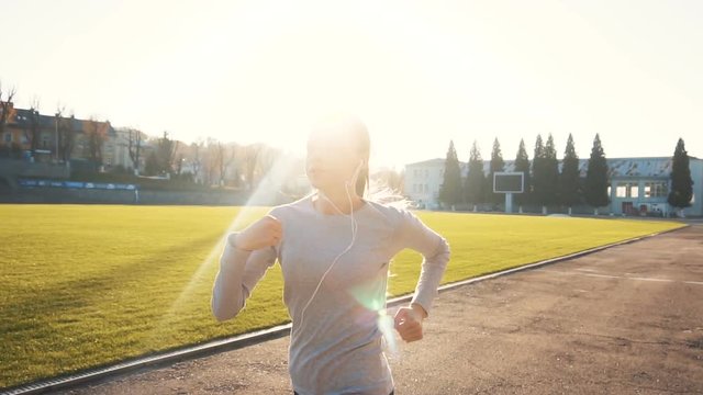 Athletic girl, in grey top and black tracksuit bottoms, is jogging in the stadium, outside slowmotion on sunny day