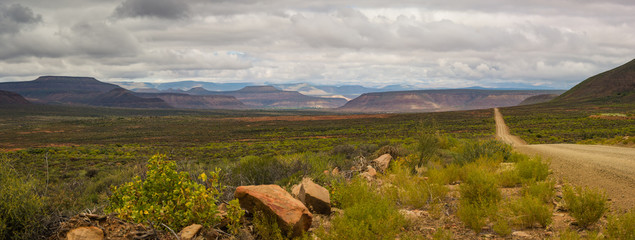 A Karoo country road in South Africa stretches out to the horizon with mountains and clouds in the background 