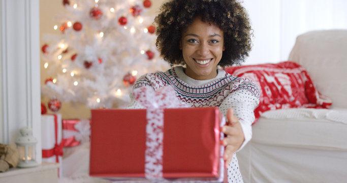 Smiling friendly woman offering a large decorative red Christmas gift to the camera with a lovely warm friendly smile in a festive living room with tree.