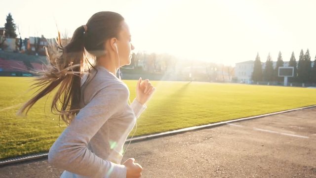Caucasian fit girl, in grey top and white headphones, running outside in the stadium during sunset