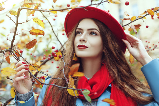 Close up outdoor portrait of young beautiful happy smiling girl wearing red hat and scarf posing near autumn tree. Model with red lips, long hair. Lady looking up.
