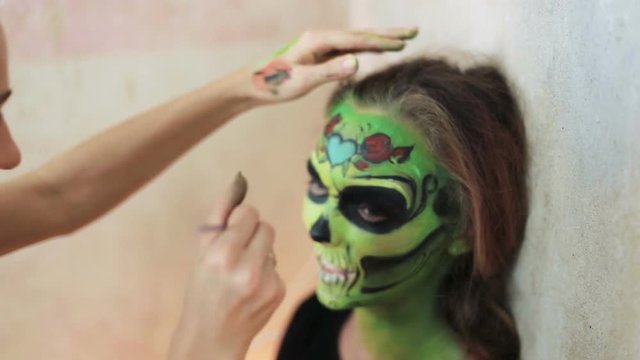 woman doing aquagrim face art on halloween make-up with her hands tassels green scary glamorous skeleton. Mexican Princess Sugar Skull.
