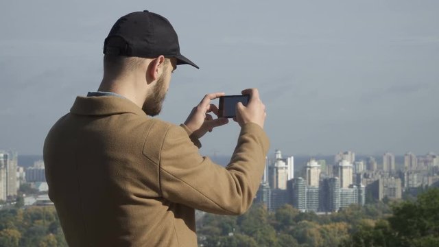 Young man in cap makes a panorama photo of city at smartphone