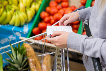 Woman with notebook in grocery store choosing vegetables, holding shopping list.