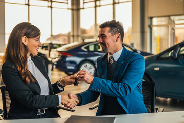 Salesman giving car keys while shaking hand of a woman in a car salon.