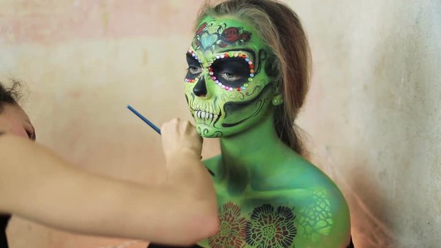 woman artist doing aquagrim face art on halloween make-up with her hands tassels green scary glamorous rhinestones skeleton. Mexican Princess Sugar Skull.