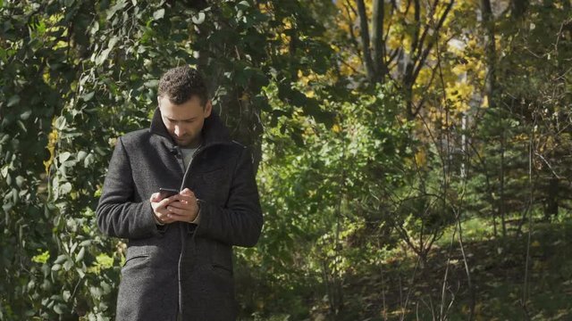 Young man in coat uses phone in autumn park