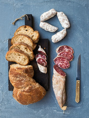 Wine snack set. Italian slami sausages and rustic bread on dark wooden board over a rough grey-blue concrete background, top view