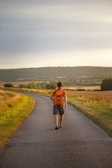 a young tourist guy walks along an asphalt road along a field in the countryside with an orange backpack and a camera around his neck in shorts and sandals in france in a burguidi in the summer