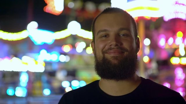 caucasian bearded guy smiling posing at camera with attractions background spending time hanging out in amusement park slowmotion closeup