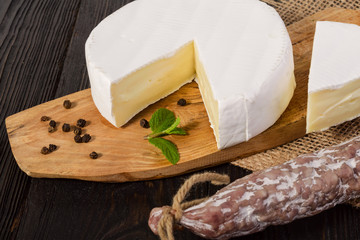 Round Camembert cheese, sliced, on a wooden board and fuet sausage, on a wooden board, with sack.