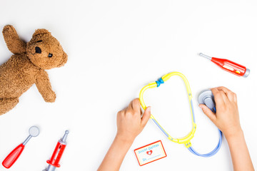 Kid hands with toy stethoscope, teddy bear and toy medicine tools on a white background. Top view.