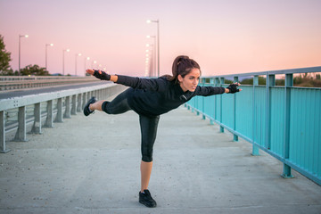 Sporty girl doing balance exercise standing on one leg with raised hands outdoors.