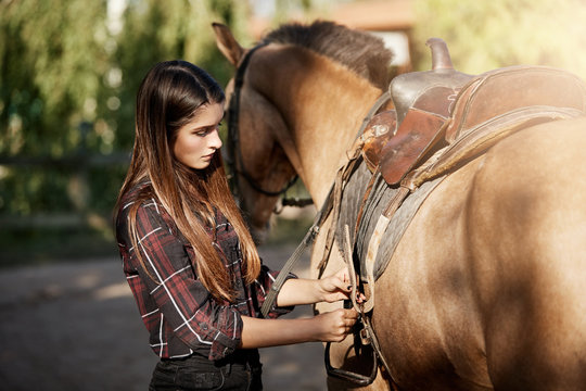 Young woman preparing to ride a horse tying down the saddle.