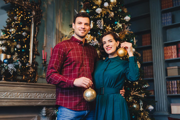 Handsome man with stubble embraces his beautiful wife, hold glass balls in hands, decorate New Year tree together, prepare for holidays. Smiling young beautiful female and her husband at home