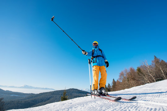 Professional skier standing on top of a mountain on a beautiful sunny winter day taking a selfie with action camera on selfie stick at the winter resort copyspace modern technologies memories concept