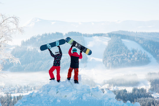 Two people stand with their backs on a snowdrift and raise their snowboards up against a white haze of snow-capped mountains and forests at winter ski resort