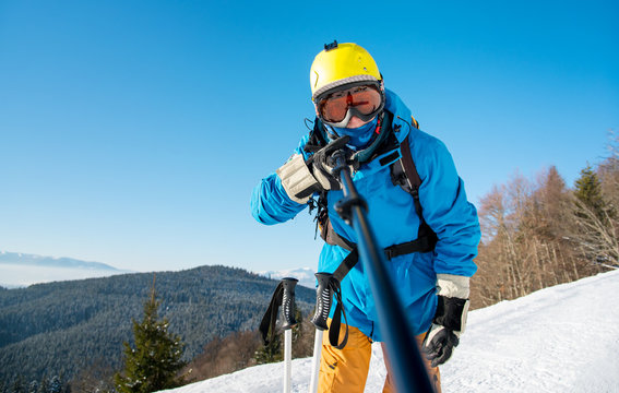 Shot of a man skier wearing ski equipment taking a selfie using monopod at the winter resort. Blue sky and winter forest on the background technology youth recreation activity