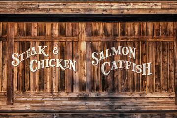 Weathered wooden shed with vertical running timber with the painted words STEAK, CHICKEN, SALMON, and CATFISH painted on in white