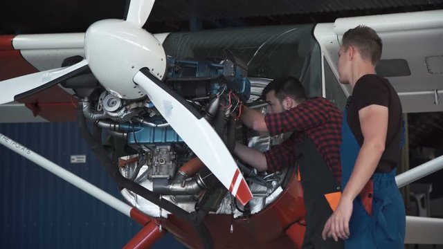 Two mechanics working on a small aircraft in a hangar with the cowling off the engine as they perform a service or repair