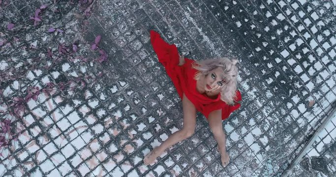 Aerial view of mysterious woman in red dress with gunshot wound in her head looking into the camera while sitting in destroyed outdoor swimming pool of abandoned building