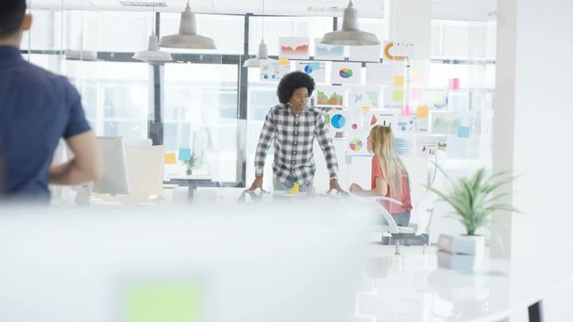  Young woman working on computer in creative office with colleagues talking in background. Shot on RED Epic.