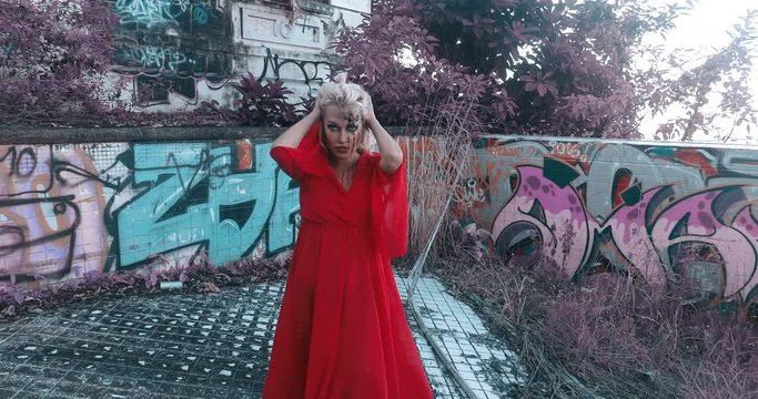 Mysterious woman in red dress with gunshot wound in her head looking into the camera while walking in destroyed outdoor swimming pool of abandoned building