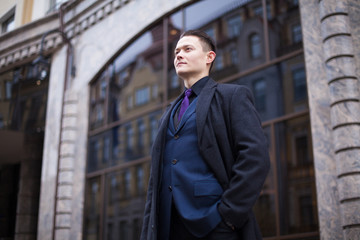 A young and purposeful banker in a blue suit and coat looks forward against the background of the bank building.