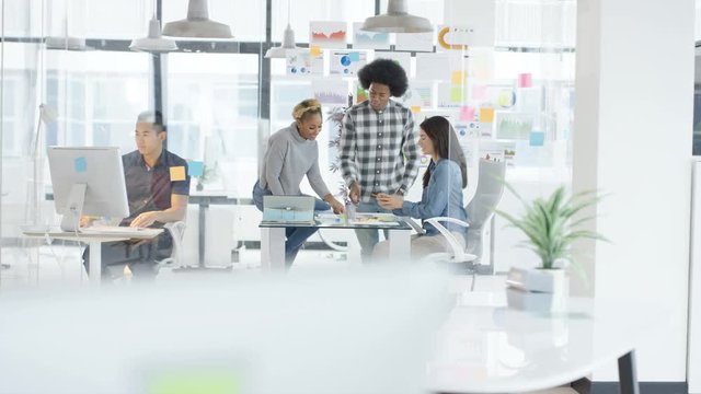  Young casual business group in a meeting in creative office with woman working on computer in foreground. Shot on RED Epic.