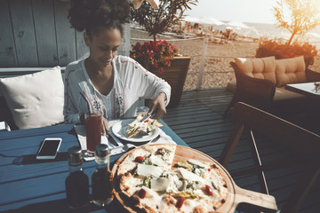 Fototapeta na wymiar Cute cheerful young black woman is sitting in street cafe with beach resort and sea in defocused background and having lunch: putting on a plate piece of delicious freshly baked pizza