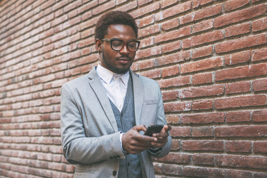 Cute black businessman sending text message with his mobile phone in front a brick wall.