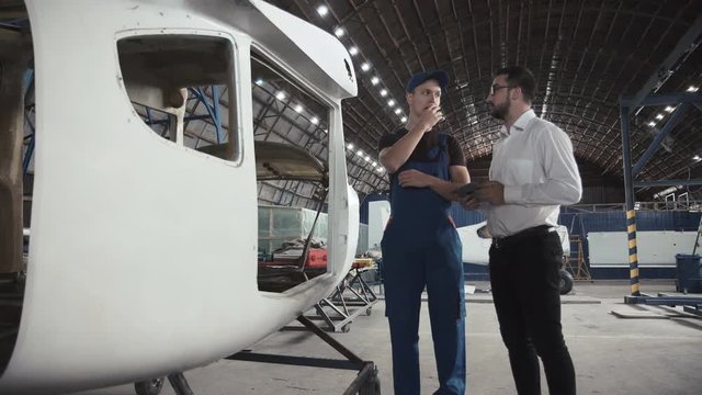 Aircraft mechanic and draftsman standing near plane carcass and talking with each other using digital tablet.