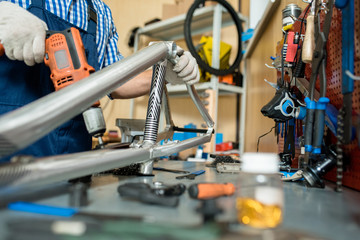 Close-up shot of unrecognizable mechanic wearing checked shirt and overall fixing bicycle frame while standing at desk