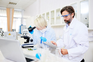 Scientist in whitecoat making notes while his colleague studying chemical substance in microscope