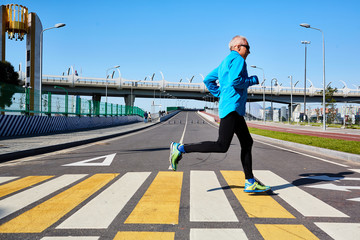 Profile view of talented senior athlete in sportswear crossing road while having running training outdoors, cityscape on background
