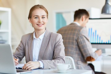 Happy businesswoman making notes in working diary with her colleague on background