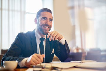 Waist-up portrait of cheerful bearded entrepreneur looking away with toothy smile while working on...