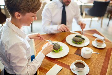 Profile view of smiling pretty businesswoman enjoying delicious burger while having lunch at modern cafe, her male colleague sitting next to her