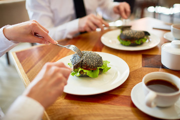 Close-up shot of unrecognizable coworkers in formalwear sitting at cafe table and enjoying appetizing burgers and herbal tea