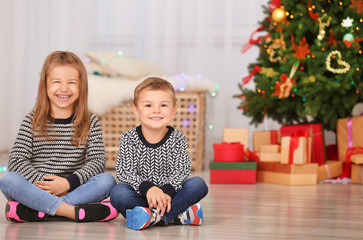 Cute little children sitting on floor in room decorated for Christmas