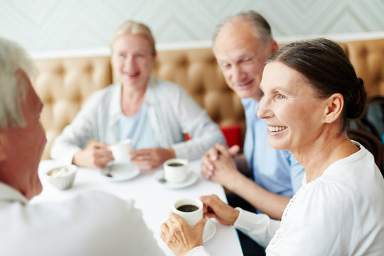 Group of senior companions sitting by table in cafe and having talk by cup of coffee