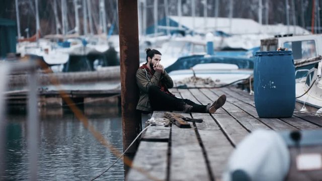  Rugged hipster man sitting alone on jetty in marina & playing harmonica