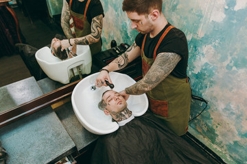 Man getting trendy haircut at barber shop. Male hairstylist in tattoos serving client, washing head