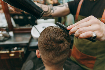Close up shot of man getting trendy haircut at barber shop. Male hairstylist in tattoos serving client, drying hair with a hairdryer
