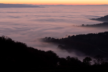 Aerial view of a valley filled by fog at sunset, with beautiful warm colors of the sky reflecting on the mist