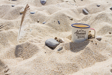 Enameled mug with clean drinking water from Lake Baikal on the shore of the lake and brown seagull feather in the sand among the stones in the summer.