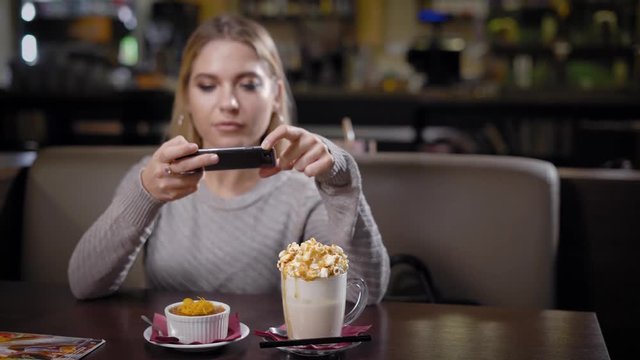 joyful young woman with blonde hair is taking photo of her cake and coffee by phone, sitting in cozy coffee house