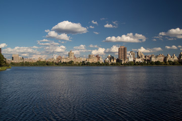 Naklejka premium Urban city landscape of buildings and skyscrapers behind the lake or river and trees, with reflection of buildings in water and clouds on blue sky